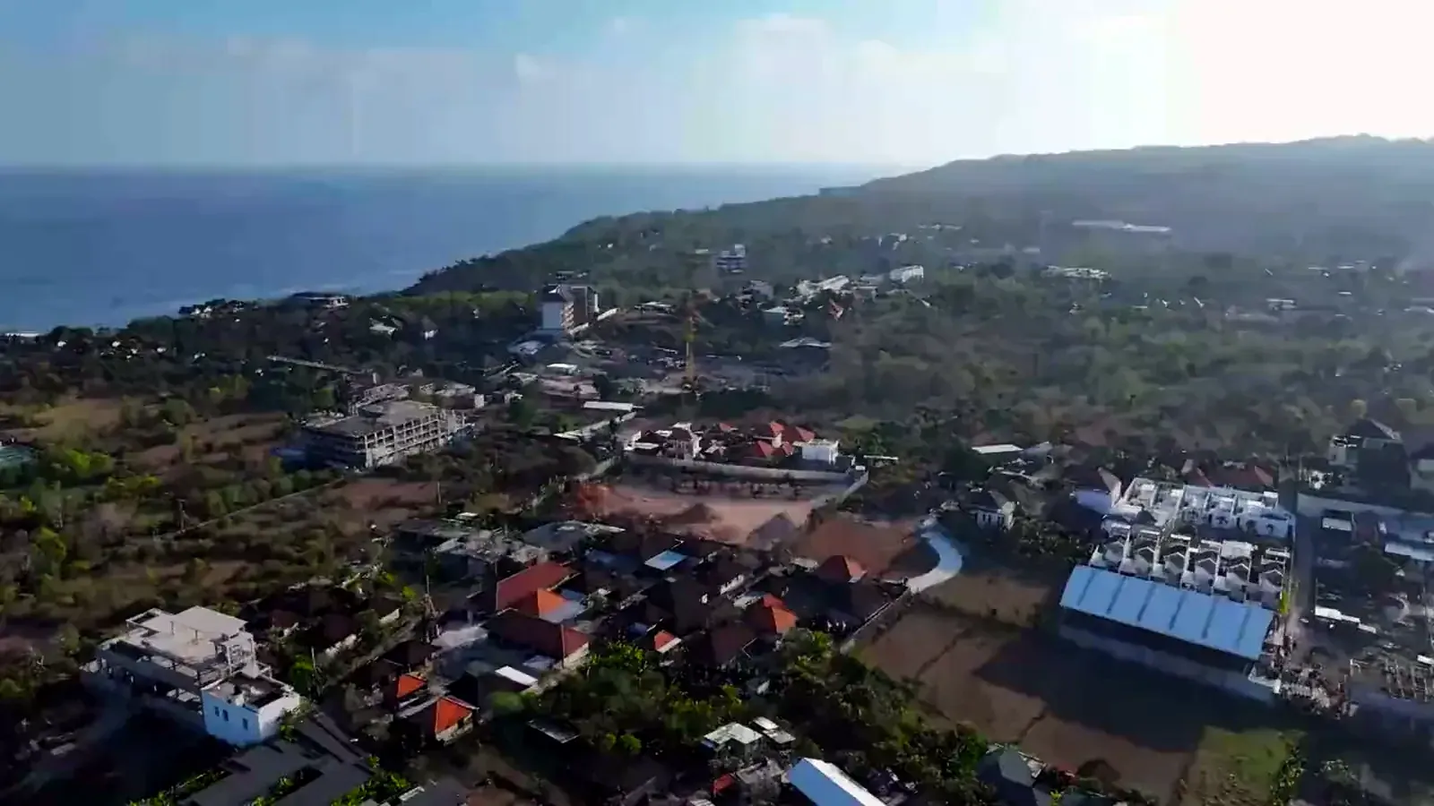 Aerial view over Ungasan on the Bukit plateau: terracotta roofs, new buildings and a construction crane among scrub, with the Indian Ocean coastline running to the horizon